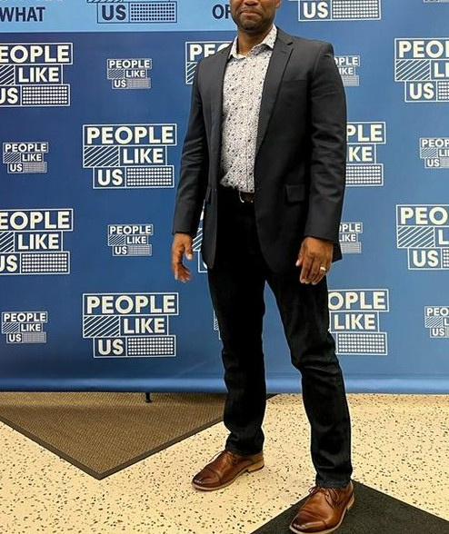 a person in a suit stands in front of a people like us banner